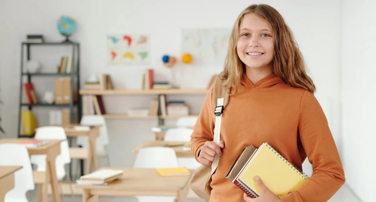 Uczennica z zeszytami i plecakiem stojąca w jasnej sali lekcyjnej z ławkami i materiałami edukacyjnymi w tle. Kadr podkreśla pozytywne nastawienie do nauki i przygotowania do egzaminu ósmoklasisty w Krakowie w ramach kursów prowadzonych w kameralnych grupach. Więcej informacji o ofercie można znaleźć na stronie Symposio.